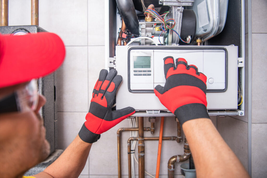 A man with gloves and a red hat works on a gas furnace